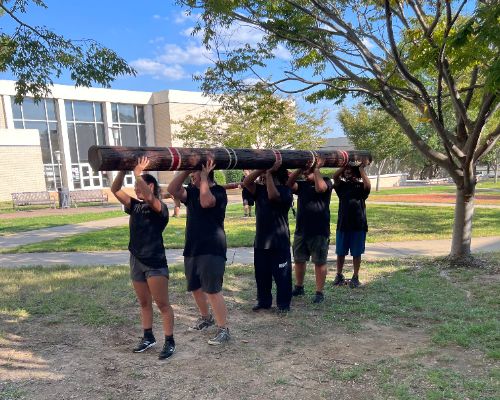  Students in athletic gear hold log above their heads.